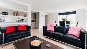 A modern living room with two black leather sofas, each with a red cushion, a coffee table with a fruit bowl, and a white shelving unit with various items. - Luxury 2 Bed/2 Shower Room Apartment in Bath City (Bath)