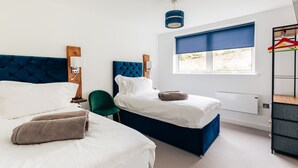 Une chambre d’hôtel avec deux lits, une fenêtre avec un store bleu, un luminaire suspendu et un porte-manteau avec des cintres colorés.