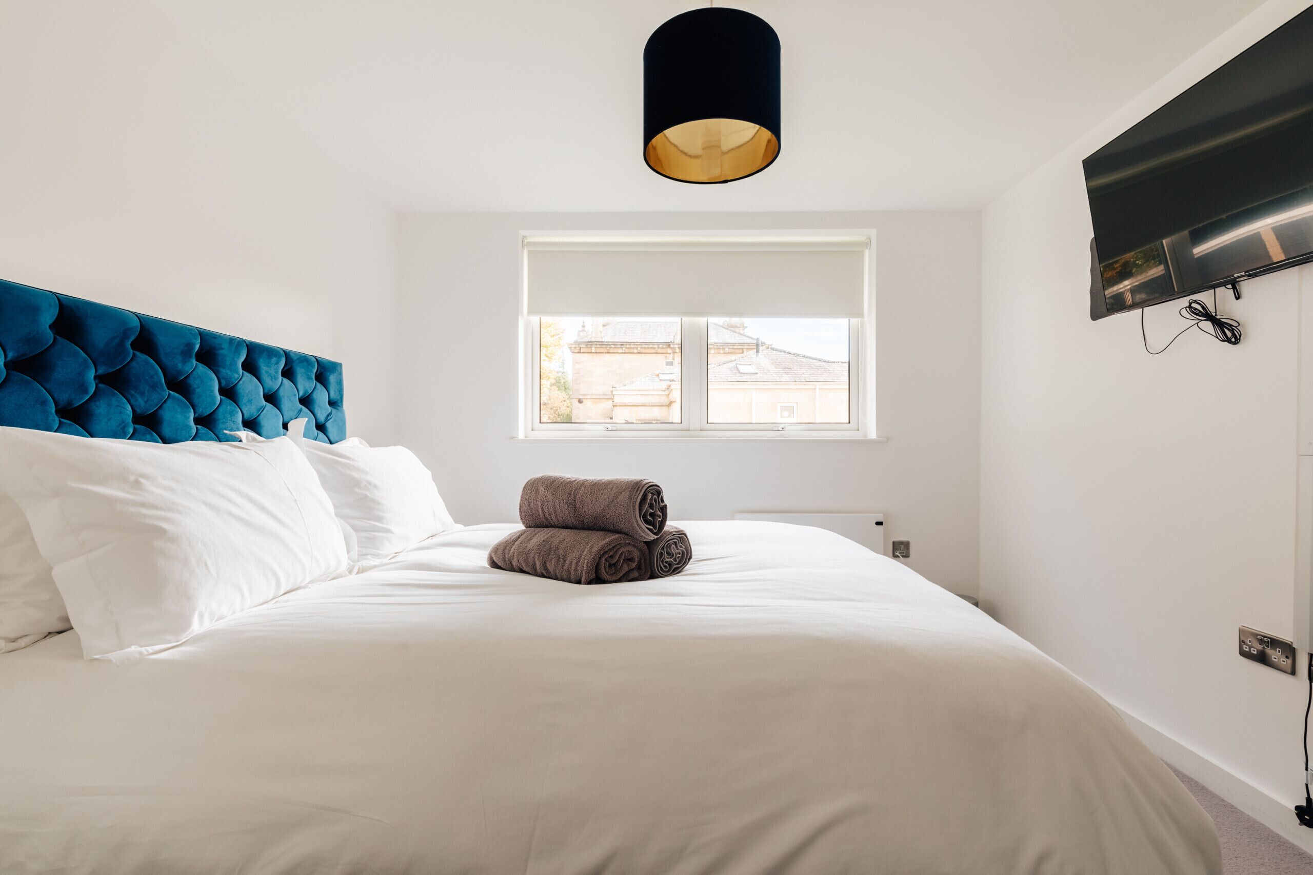 Una cama bien hecha con ropa de cama blanca y marrón, un cabecero capitonado azul y una ventana con vistas a edificios.
