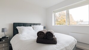 Ein ordentlich bezogenes Bett mit weißer und dunkelgrauer Bettwäsche, ein Nachttisch mit Lampe und ein Fenster mit Blick auf Bäume und Gebäude.