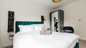 A neatly made bed with white linens and rolled towels, a green upholstered headboard, and a wall-mounted lamp.