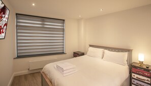 A neatly made bed with white linens, a window with horizontal blinds, a bedside table with a lamp, and a wall-mounted artwork. - Delightful Modern Apartment in Old Town Swindon (Swindon)