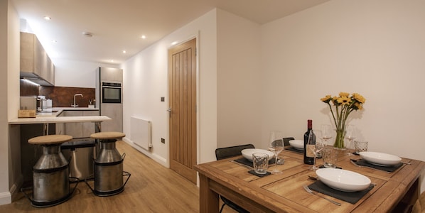 A modern kitchen with a wooden dining table set for four, a coffee machine, and a vase with flowers.