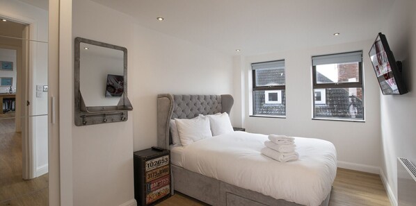 A bedroom with a large bed, a headboard, a dresser, a mirror, and a television on the wall.