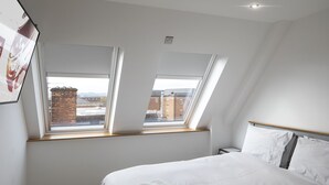 A bedroom with a large bed, two skylights, a wooden headboard, and a view of buildings outside.