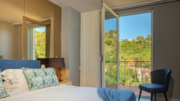 A bedroom with a bed, a chair, a nightstand, a lamp, and a balcony with a view of greenery and buildings.