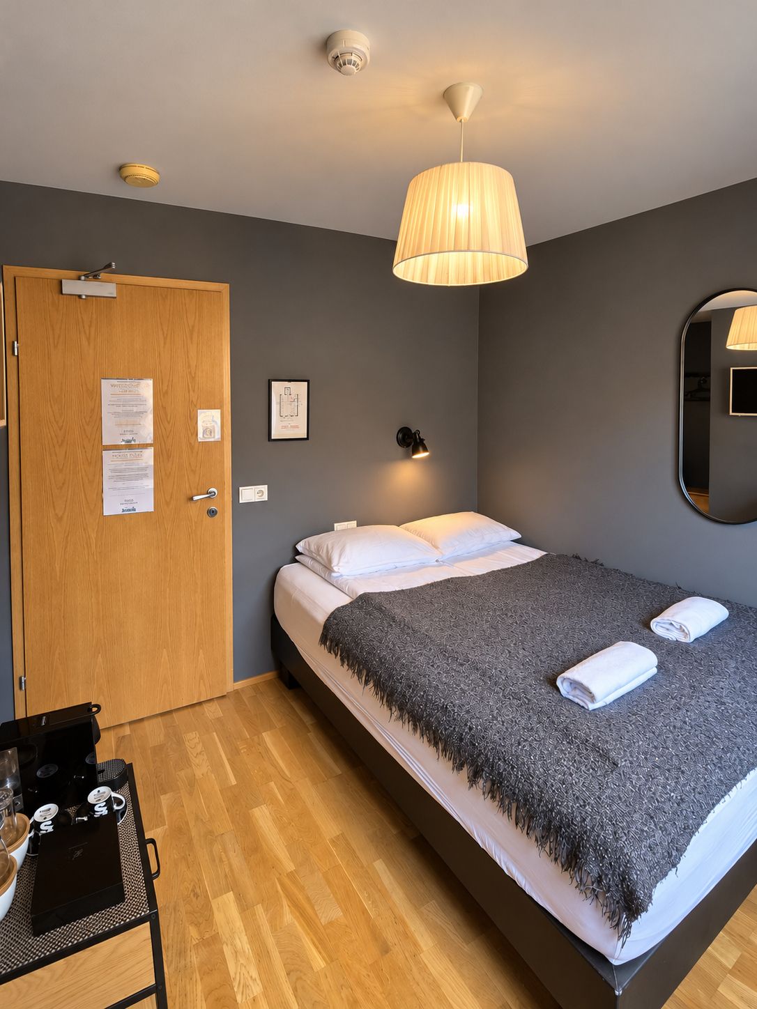 A hotel room with a bed, a wooden door, a small table with a kettle, a mirror, and a ceiling light fixture.