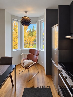 A modern kitchen with a built-in oven, a dining table with chairs, a large window with autumn foliage view, and a unique ceiling light fixture. - Torg Guest House (Akureyri)