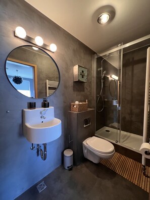Un baño moderno con un espejo redondo, un lavabo blanco, un inodoro y una ducha con mampara de vidrio.