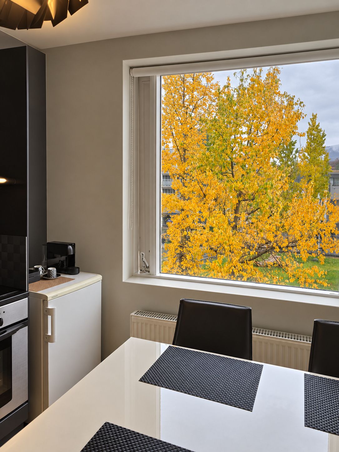 Una cocina moderna con zona de comedor, un ventanal con vistas a follaje amarillo y una cafetera.