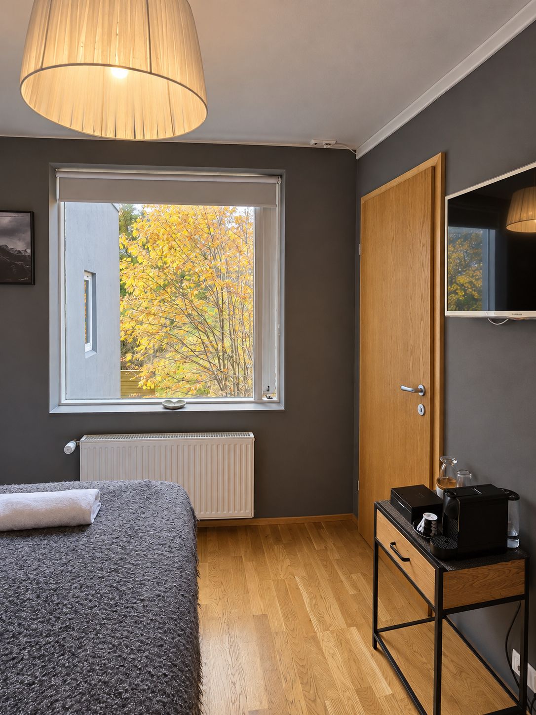 Un dormitorio con cama, ventana con vistas a árboles otoñales, radiador, puerta de madera y una pequeña mesa con objetos encima.