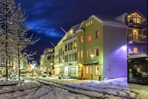 Calle nevada de noche con edificios, un escaparate visible y una vía de tranvía.