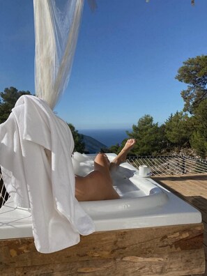 A person relaxing on a white outdoor bathtub with a view of the sea and trees.