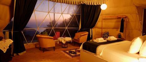 A tent with a bed, two chairs, a small table, and a window with curtains.