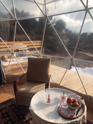 A cozy room with a large glass window, a comfortable chair, and a table with a fruit bowl and candles. - Türkdomes Glamping (Fethiye)