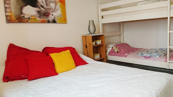 Une chambre avec un lit superposé, un lit simple avec des oreillers rouges et jaunes, une petite armoire en bois et un tableau encadré accroché au mur.