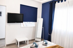 Ein Hotelzimmer mit Flachbildfernseher, einer kleinen weißen Bank, einem Bett mit blumigem Überwurf, einem großen Spiegel und einem Fenster mit blauer Jalousie.