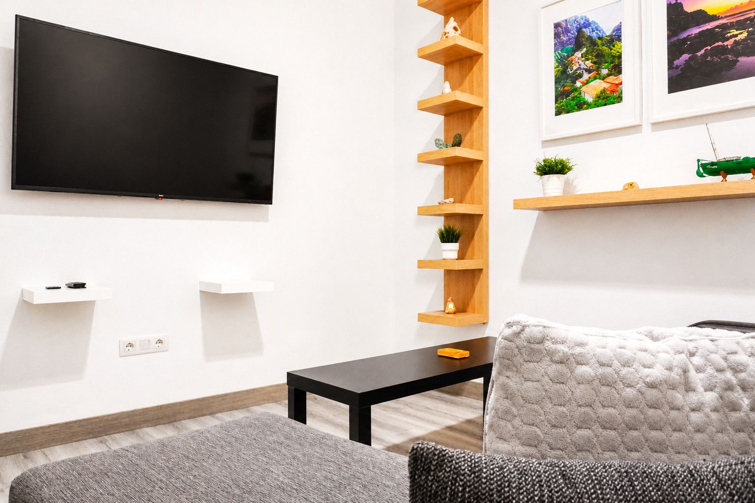 Un salon moderne avec un téléviseur à écran plat fixé au mur, une table basse noire, un canapé gris et un fauteuil blanc. On y trouve des étagères en bois ornées d’objets décoratifs et une petite plante en pot.