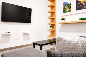 Een moderne woonkamer met een flatscreen televisie aan de muur, een zwarte salontafel, een grijze bank en een witte fauteuil. Er zijn houten planken met decoratieve objecten en een kleine plant in een pot.