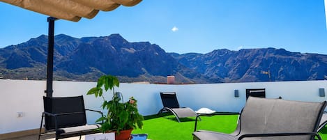A terrace with a view of mountains, a table with a plant, and two chairs.