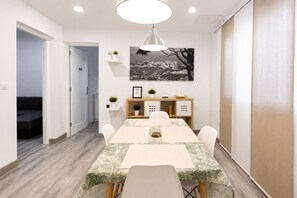 Een eetruimte met een lange tafel, stoelen, een hangende lamp, een aan de muur bevestigd schilderij en een klein kastje met planten.