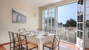 A dining area with a table set for four, chairs, a painting on the wall, and a view through large windows. - Villa 100m From the Beach (Pornic)