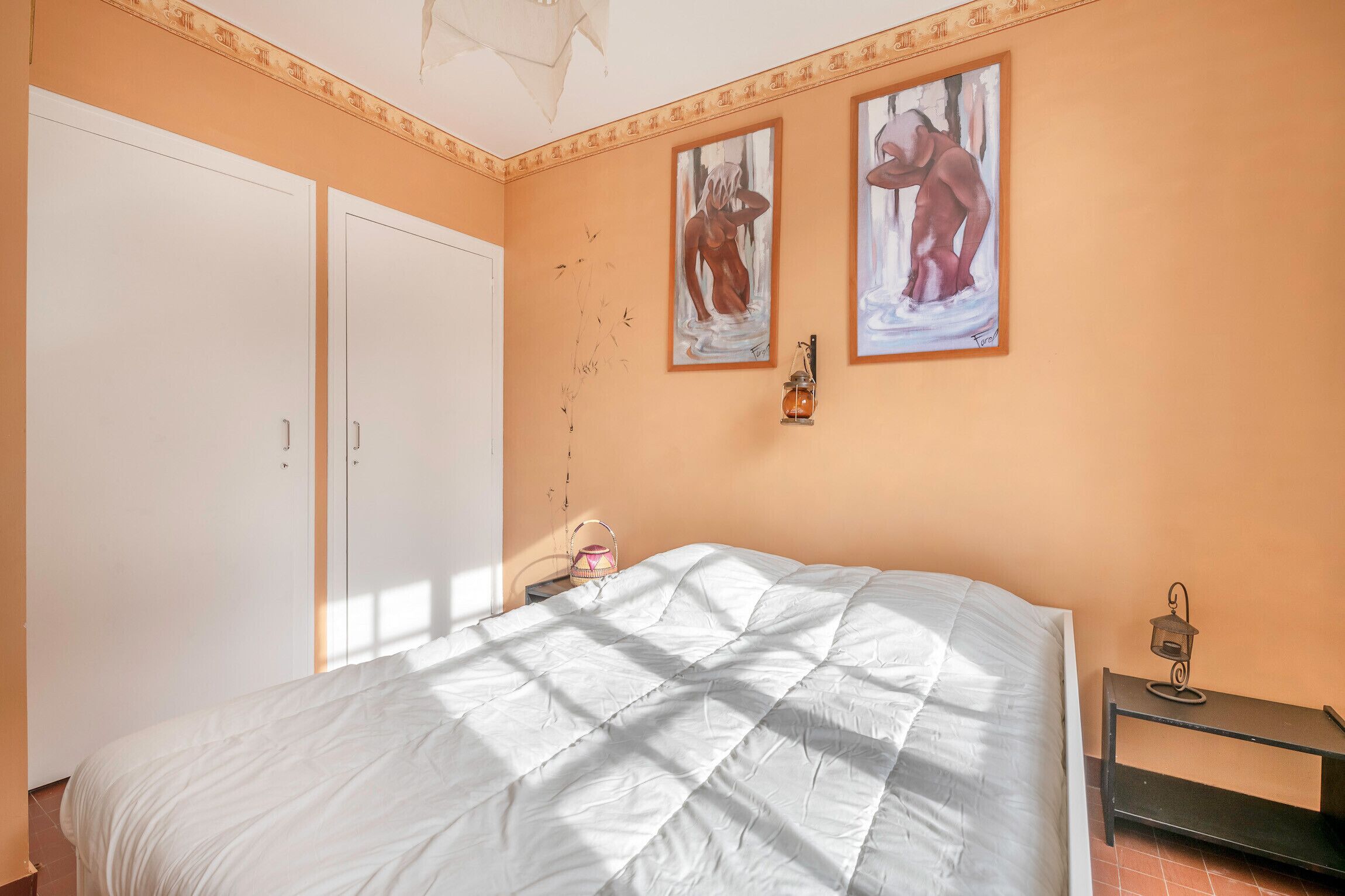 Une chambre avec un grand lit, une armoire, deux tableaux au mur, une table de chevet avec une lanterne et une petite commode.