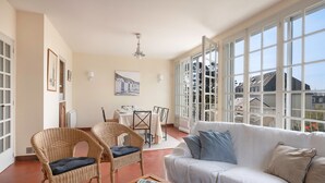 A spacious living room with a white sofa, two wicker chairs, a dining table with chairs, and large windows offering a view of buildings. - Villa 100m From the Beach (Pornic)