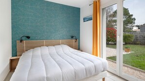 Une chambre à coucher comprenant un lit, une table de chevet, une lampe fixée au mur et une porte-fenêtre coulissante donnant sur un jardin.