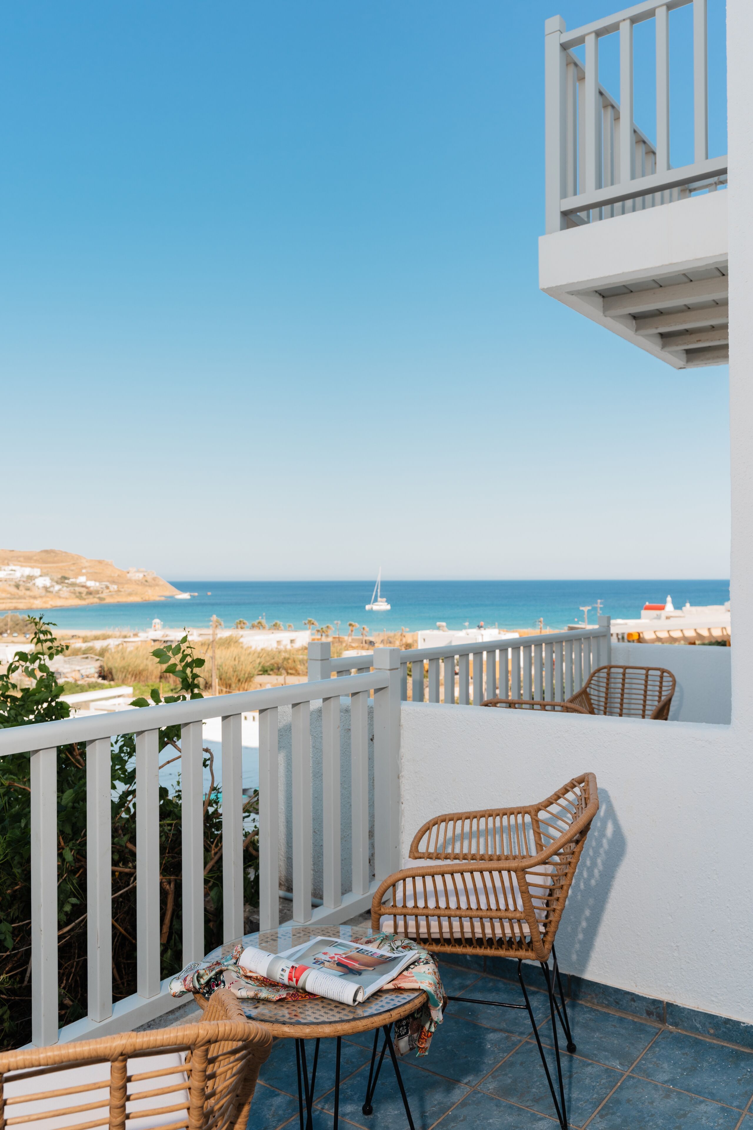 A balcony with wicker chairs and a small table, overlooking a beach with a clear sky and distant boats.