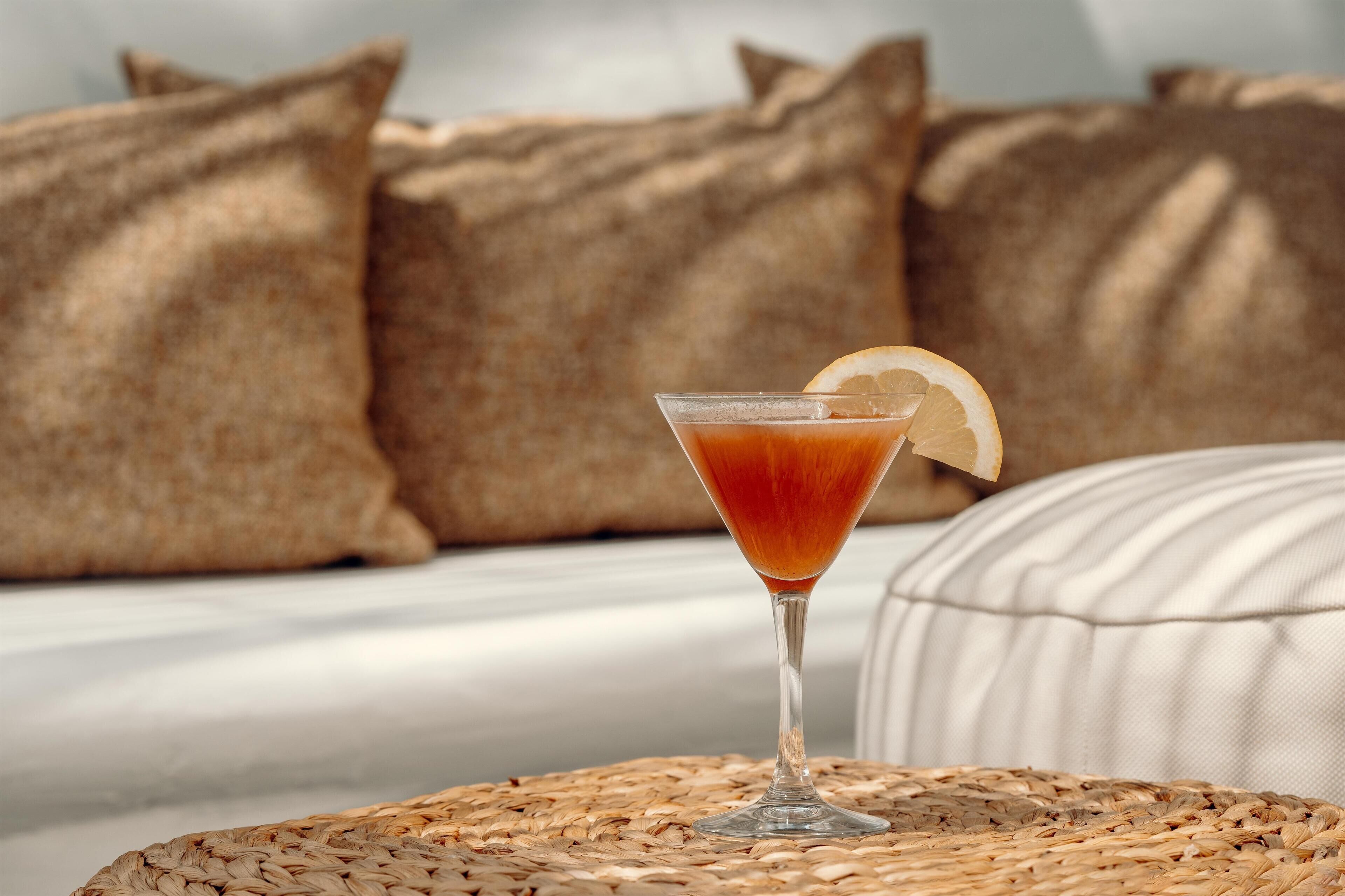 Un cocktail servi dans un verre, posé sur un dessous de verre en fibres tressées, avec un coussin et des oreillers en arrière-plan.