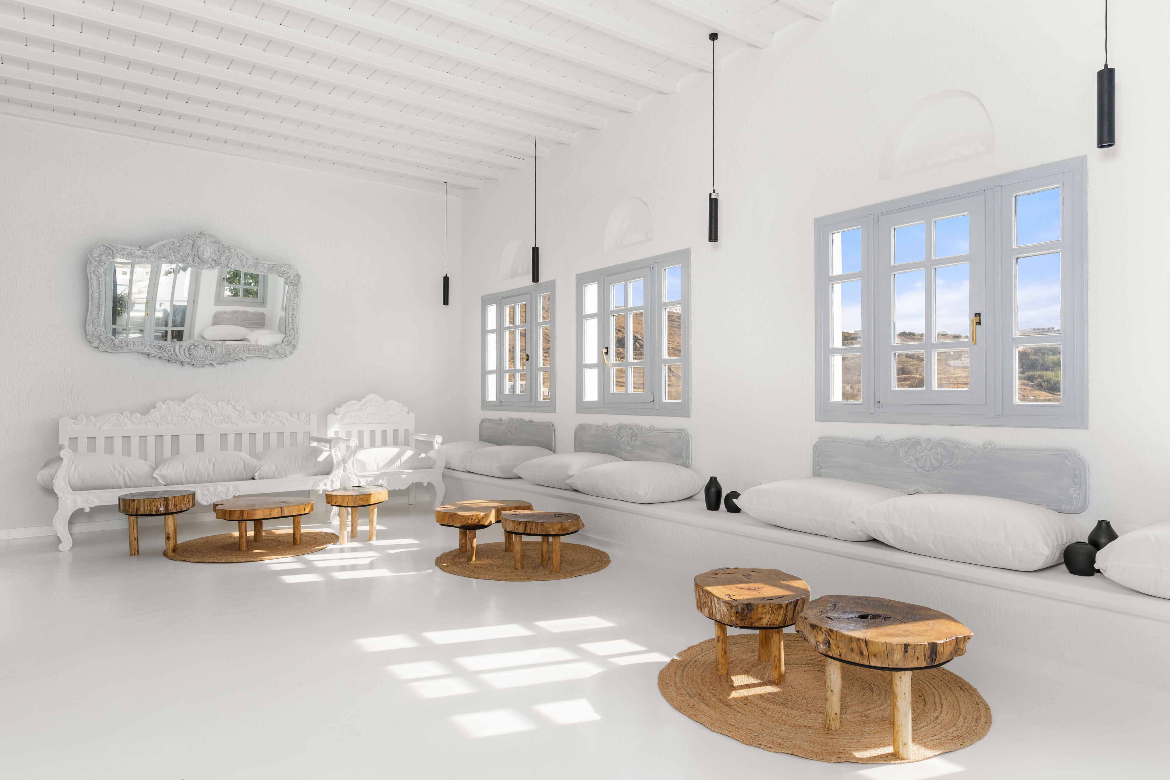 Une pièce lumineuse et blanche, avec un canapé blanc, des tables rondes en bois et de grandes fenêtres.