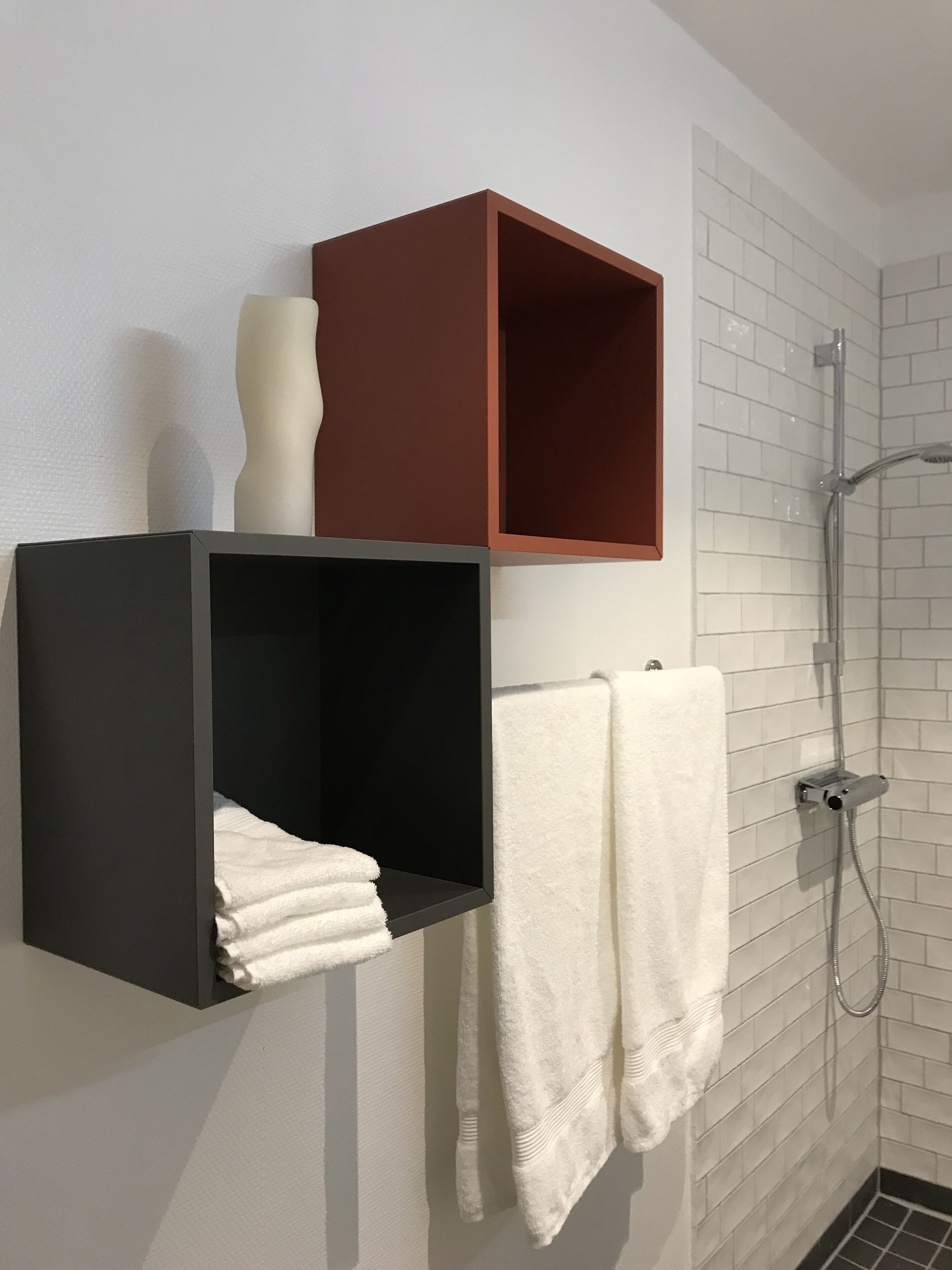 Un baño con paredes de azulejos blancos, una ducha y dos barras para toallas con toallas blancas. Hay dos unidades de almacenamiento negras y una marrón en la pared.