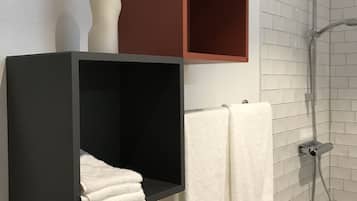 Un baño con paredes de azulejos blancos, una ducha y dos barras para toallas con toallas blancas. Hay dos unidades de almacenamiento negras y una marrón en la pared.