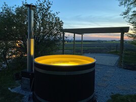 Un jacuzzi con estructura metálica y un banco de madera bajo una pérgola, situado en un jardín con vistas a campos y al atardecer.