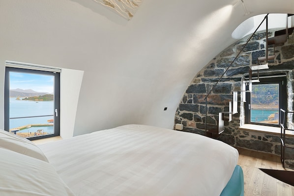 A bedroom with a bed, a large window with a view of the sea, and a stone wall.