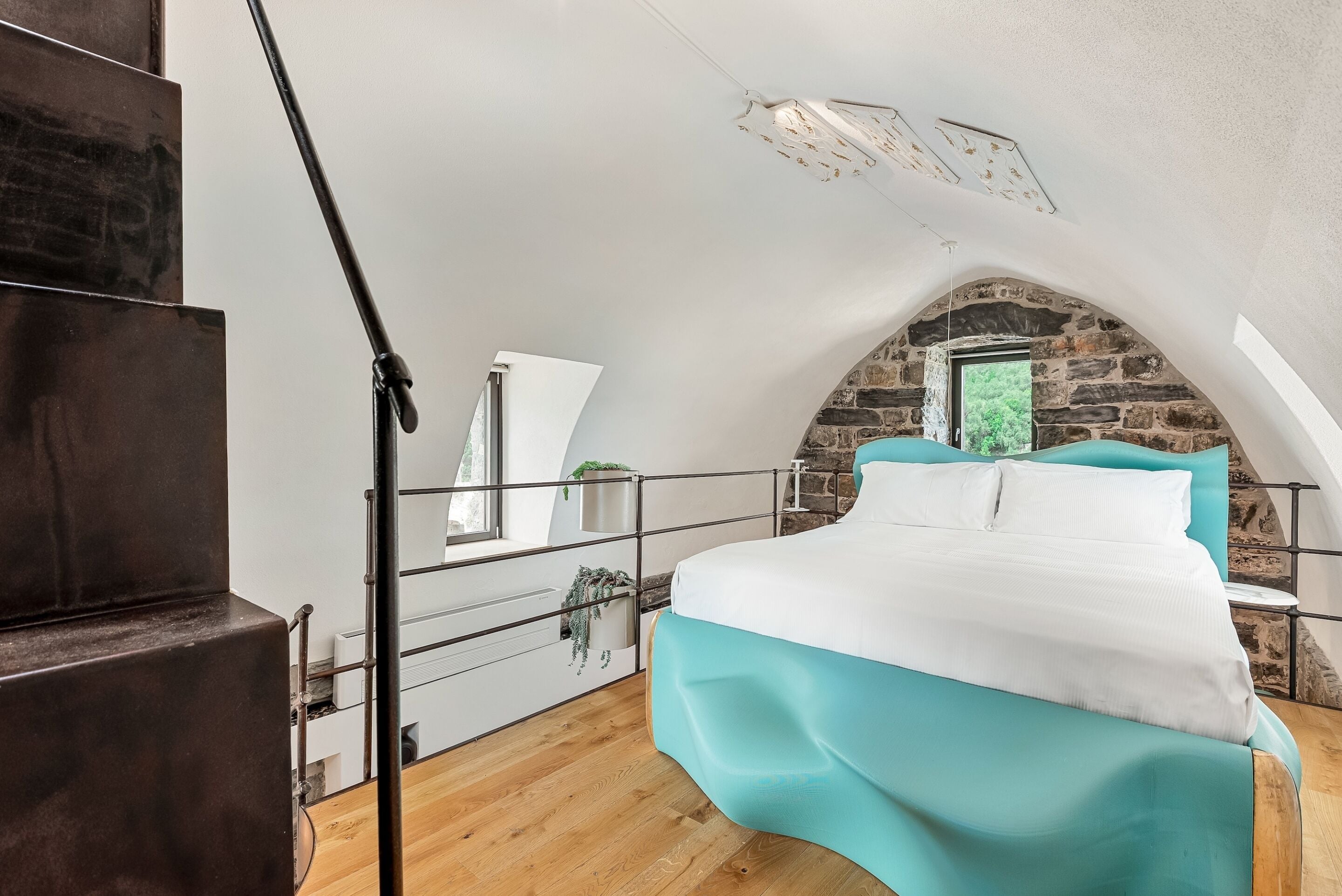 Un dormitorio con techo inclinado, una cama con ropa de cama color turquesa, un aparador blanco y una pared de piedra.