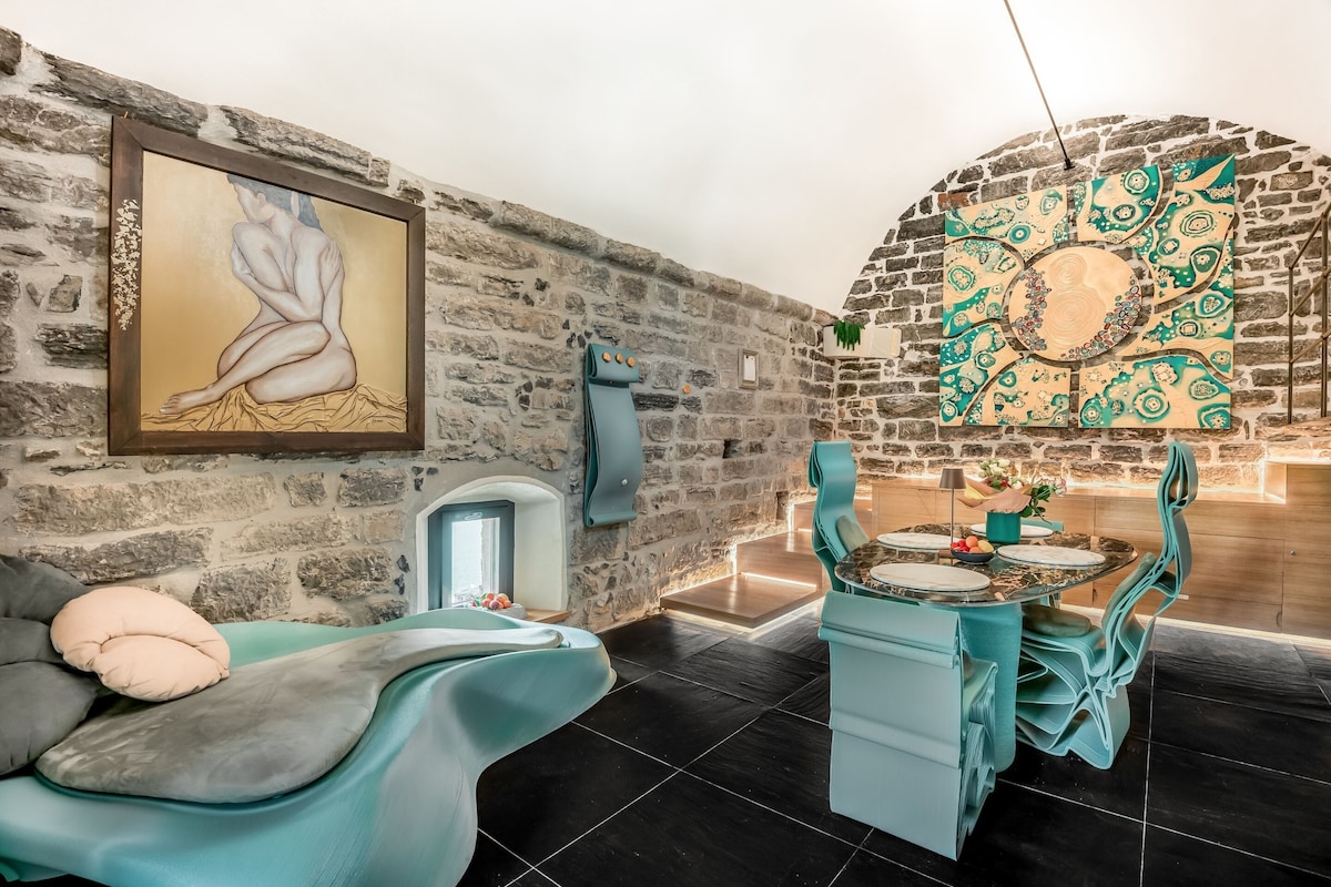 Una habitación moderna con pared de piedra, un cuadro, una chimenea, un comedor con sillas color turquesa y un sofá con cojines.