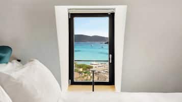 A room with a bed, a window with a view of the sea, and a chair.