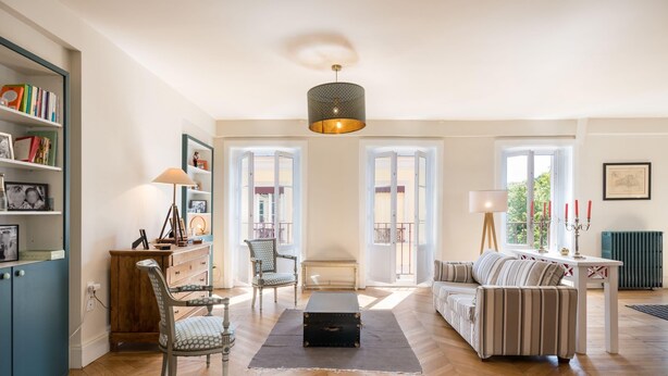 Un salon spacieux doté d’un parquet en point de Hongrie, d’un pouf noir placé au centre, d’un canapé à rayures, d’une armoire en bois et d’une bibliothèque.