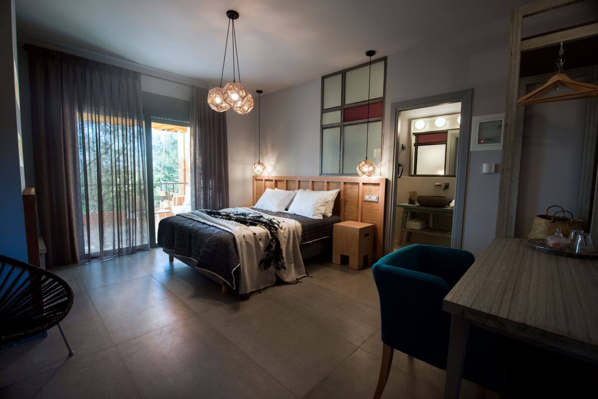 A modern hotel room with a large bed, a desk, and a chair. There is a window with curtains, a mirror, and a hanging light fixture.