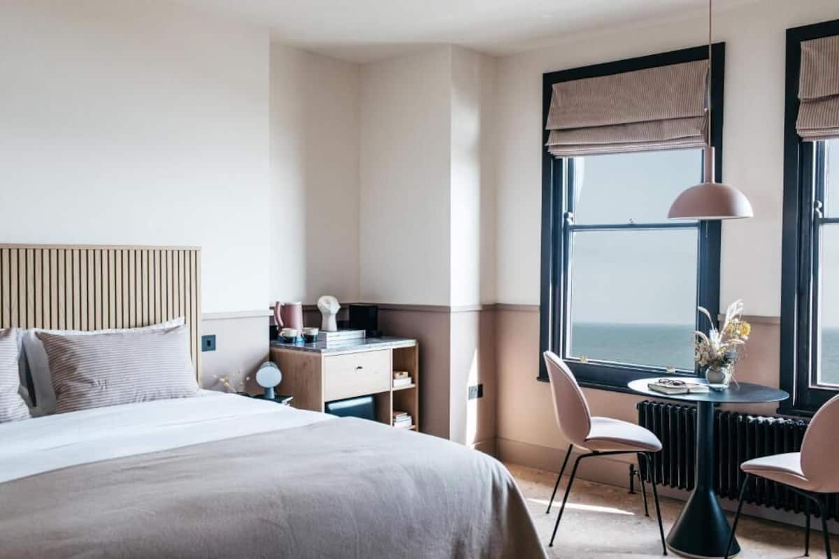 Hotellihuone, jossa on sänky, työpöytä, tuoli, maljakko pöydällä ja suuret ikkunat, joista avautuu näkymä merelle.