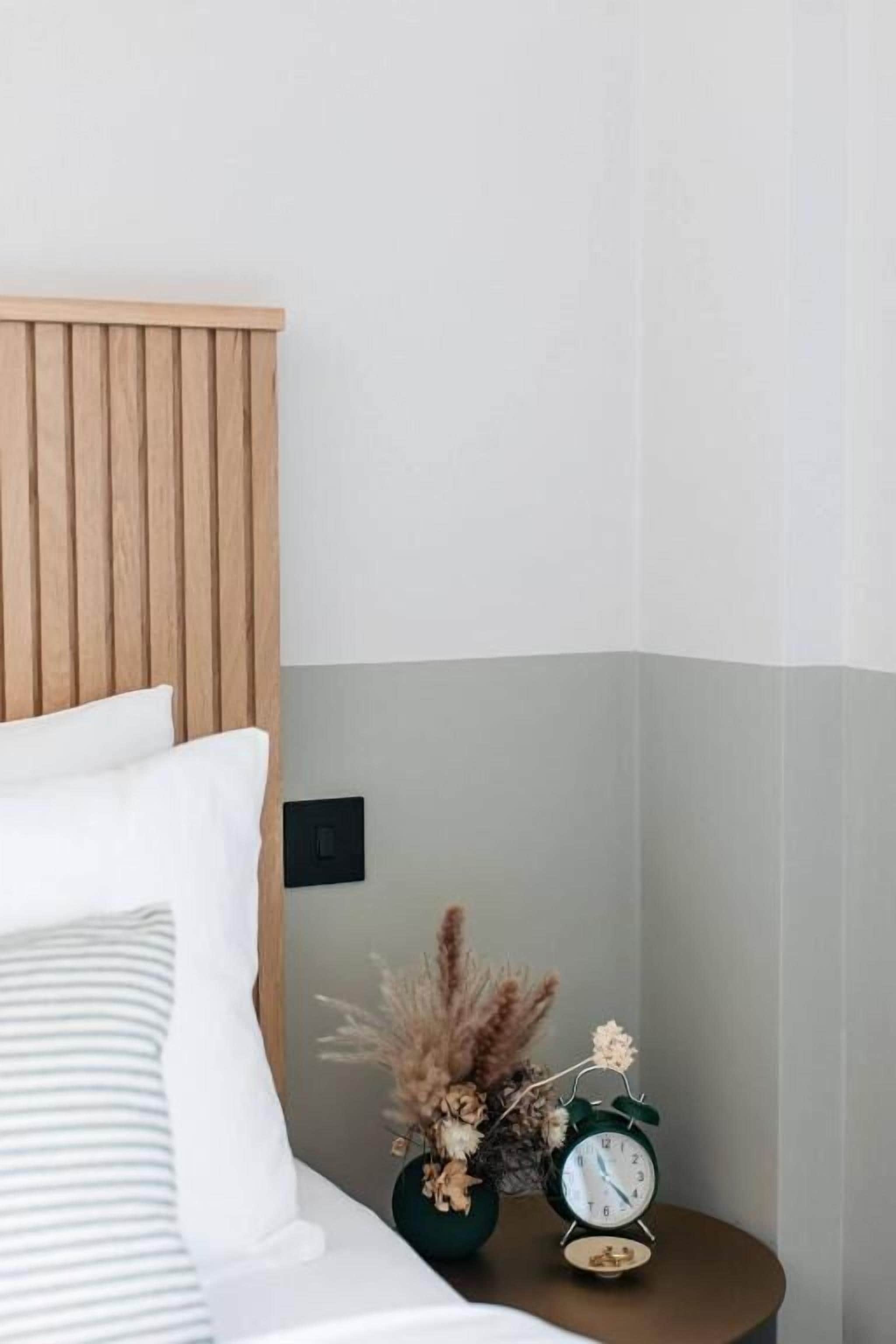 A neatly made bed with white and striped pillows, a wooden headboard, and a small bedside table with a green vase of dried flowers and a green alarm clock.