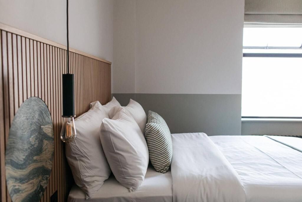 A neatly made bed with white bedding and several pillows, a wooden headboard, and a wall-mounted lamp.