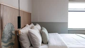 Una cama bien hecha con ropa blanca y varias almohadas, un cabecero de madera y una lámpara fijada a la pared.