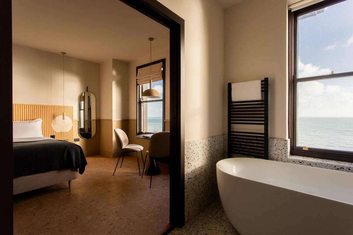 Moderni hotellihuone, jossa on suuri sänky, oleskelualue tuolilla ja pöydällä, kylpyamme ja ikkunasta avautuva merinäköala.