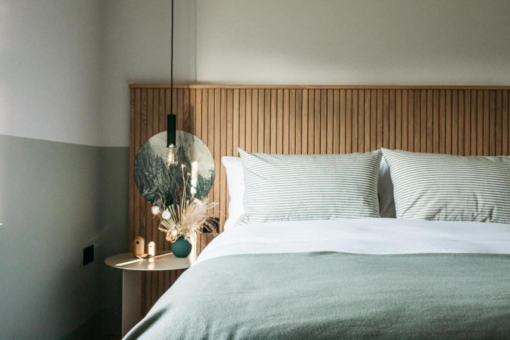 Un dormitorio ordenado con cabecera de madera, un espejo redondo y una mesita de noche con un jarrón de flores secas.