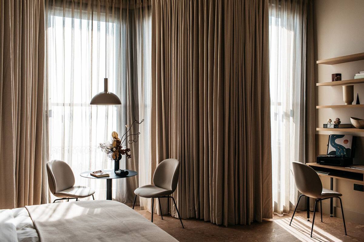 Hotellihuone, jossa on sänky, kaksi tuolia, pieni pöytä maljakolla ja työpöytä hyllyineen.