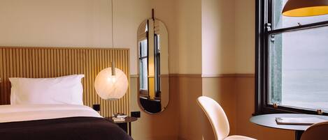 A hotel room with a bed, a round mirror, a desk with a chair, and a window with a view.