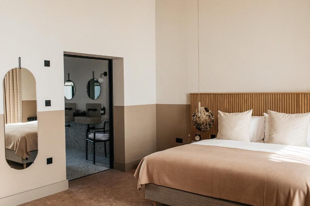 A modern hotel room with a large bed, a wooden headboard, a mirror, and a small desk with a chair.